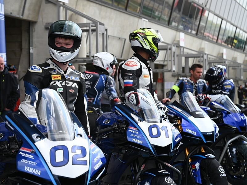 plusieurs pilotes alignés sur leurs motos Yamaha dans les stands, en tenue de course et prêts à prendre la piste.