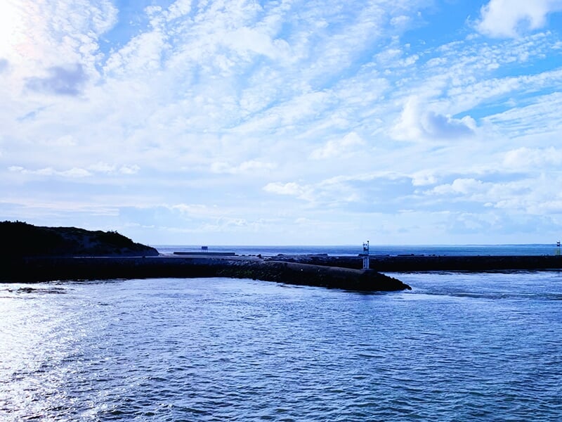 Paysage maritime avec un ciel bleu nuageux, un phare blanc sur une digue rocheuse et une étendue d'eau scintillante.