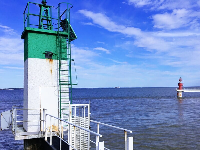 Un phare vert et blanc sur une jetée, avec un second phare rouge visible au loin et un ciel bleu clair.