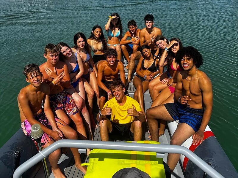 Groupe de jeunes en maillots de bain assis sur un bateau gonflable au soleil, souriants et posant pour la photo.