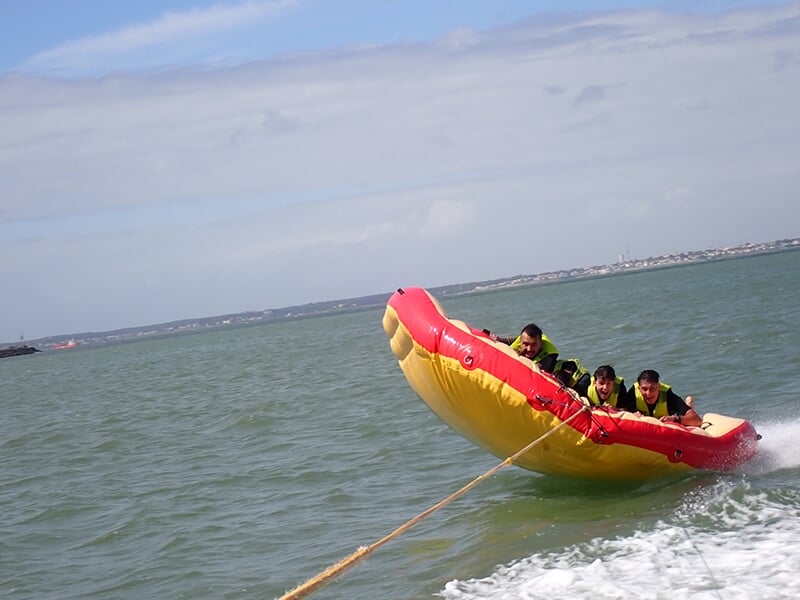 Trois personnes accrochées à une bouée gonflable rouge tirée par un bateau, rebondissant sur l'eau.