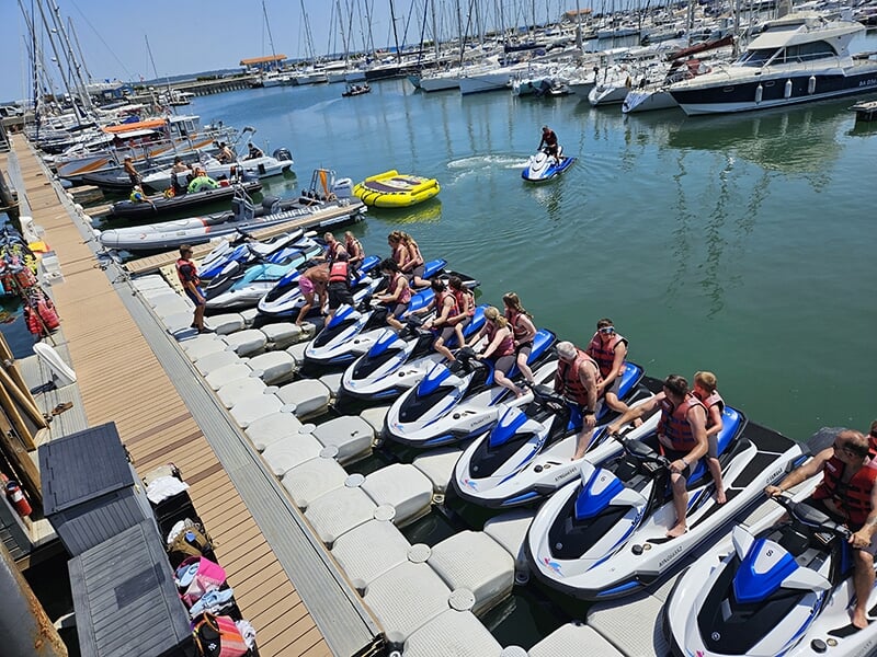 Groupe de personnes en gilets de sauvetage assis sur des jet-skis bleus et blancs, amarrés sur un quai dans un port rempli de bateaux.
