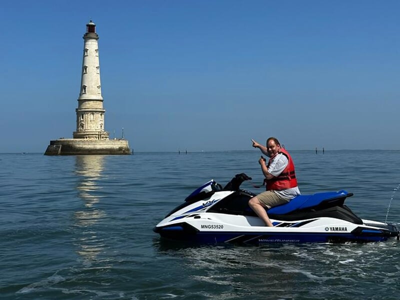 Homme en gilet de sauvetage rouge posant sur un jet-ski devant un phare en pierre, au milieu d'une mer calme sous un ciel bleu.