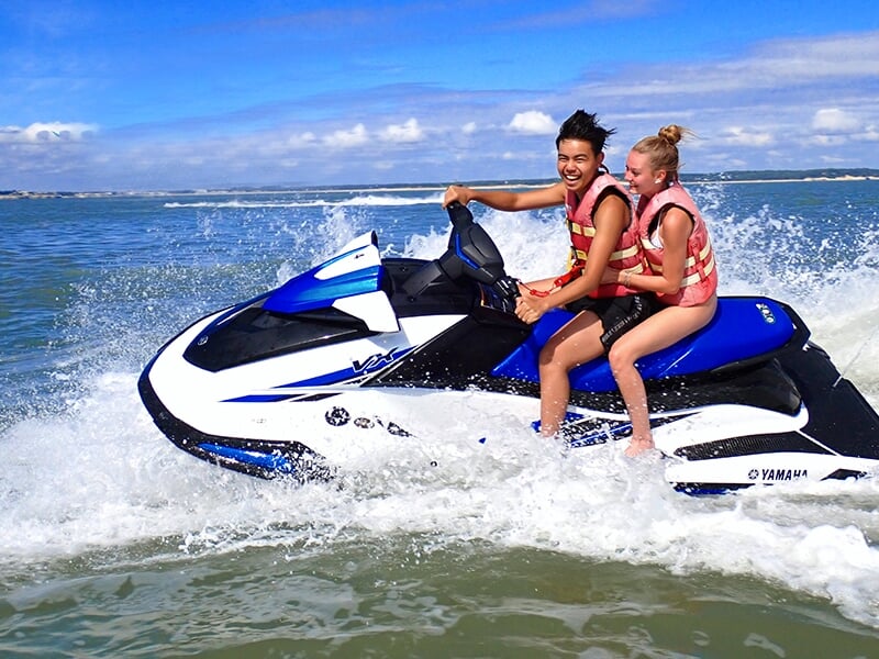 Deux personnes souriantes en gilets de sauvetage sur un jet-ski bleu et blanc, éclaboussures d'eau en arrière-plan sous un ciel ensoleillé.