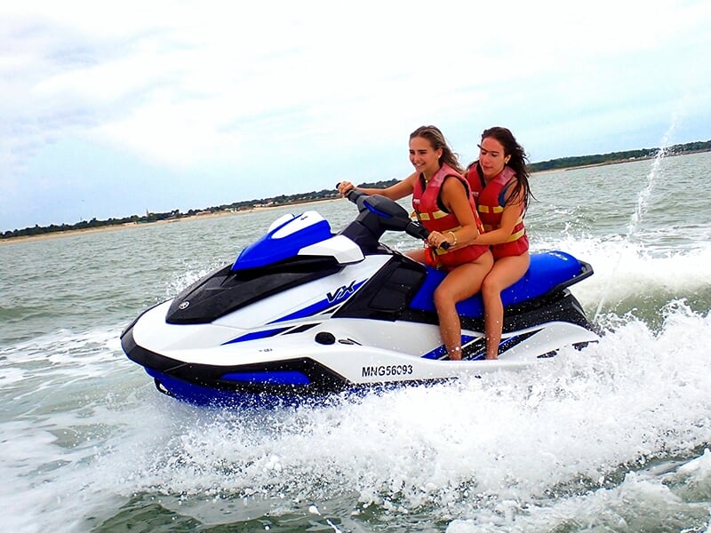 Deux femmes en gilets de sauvetage rouges conduisant un jet-ski bleu et blanc, traversant une eau légèrement agitée.