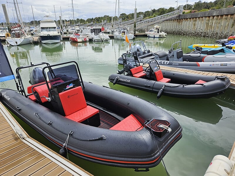 Deux semi-rigides noirs avec intérieur rouge flottant sur une eau calme dans un port, avec des voiliers à l’arrière-plan.