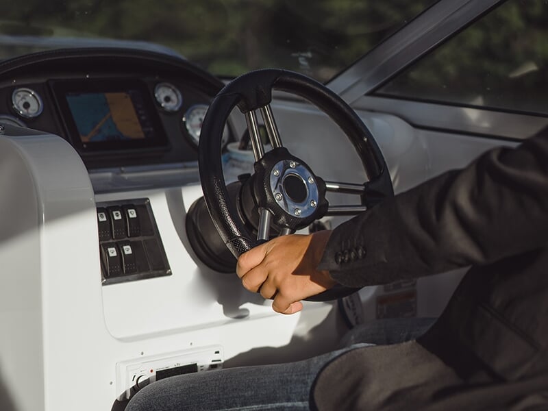 Gros plan sur les mains d'une personne tenant le volant d'un bateau, avec un tableau de bord équipé de jauges et d'un écran GPS en arrière-plan.