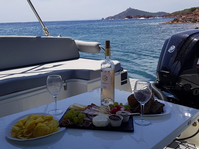 Table à bord d'un bateau avec vin rosé, verres, plateau de charcuterie et fromages, vue sur la mer bleue et une colline en arrière-plan