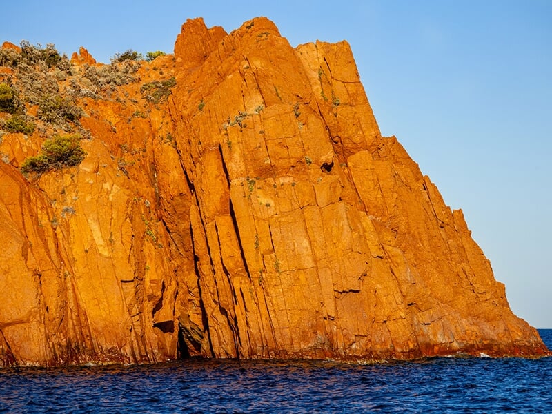 Formation rocheuse orangée surplombant la mer bleue, baignée par la lumière dorée du soleil, avec quelques arbustes verts en haut