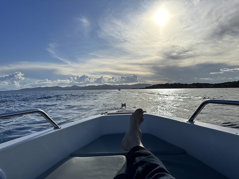 Pieds allongés sur un bateau, vue sur l'océan scintillant sous un soleil éclatant, ciel bleu parsemé de nuages et montagnes à l'horizon