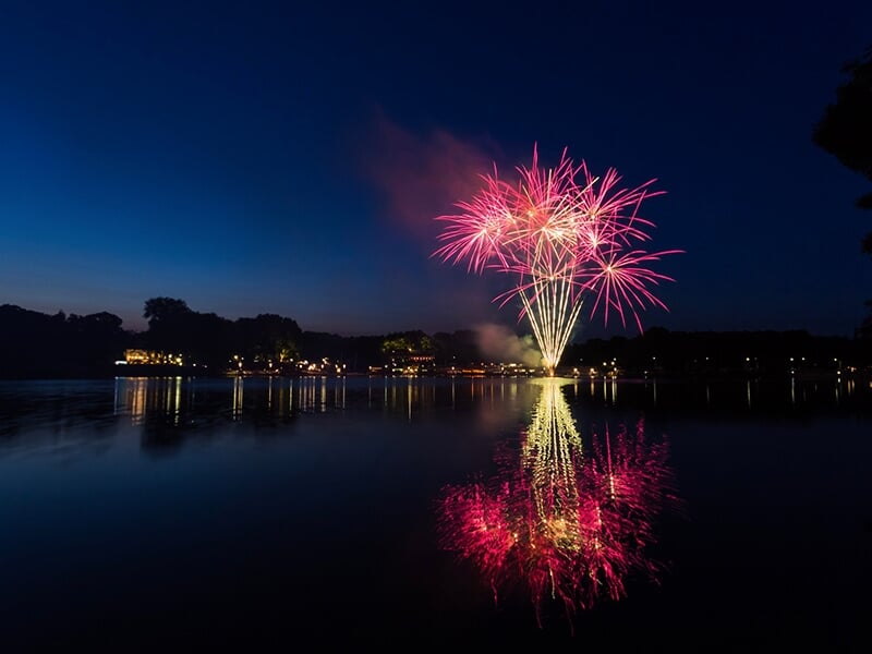 Feux d'artifice roses éclatant dans le ciel nocturne, avec leur reflet illuminant la surface calme d'un lac, entouré d'ombres d'arbres et de lumières de ville en arrière-plan