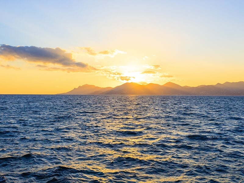 Coucher de soleil sur la mer avec des montagnes en silhouette à l'horizon, le ciel teinté de jaune et d'orange reflétant sur les vagues.