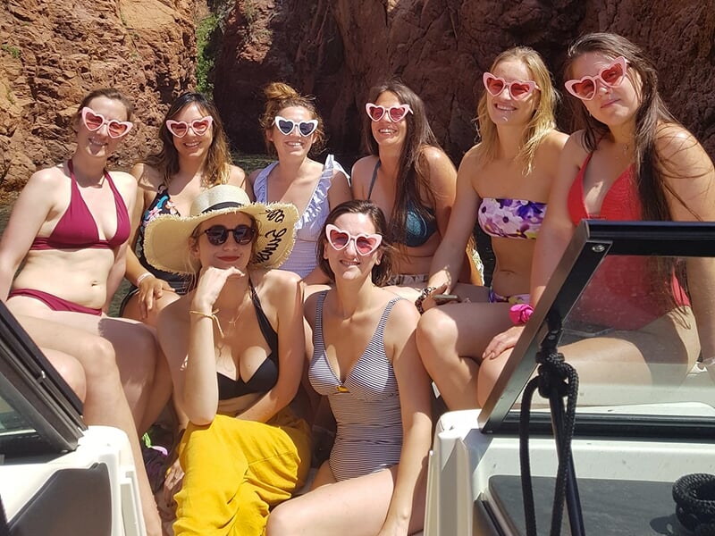Groupe d'amies portant des lunettes en forme de cœur et des tenues de plage, posant sur un bateau dans un décor rocheux naturel.