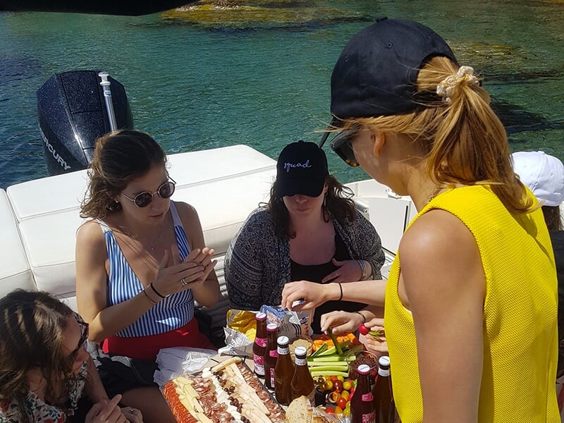 Groupe de personnes partageant un pique-nique sur un bateau, avec une variété de nourritures et de boissons, amarré près d'eaux turquoise.