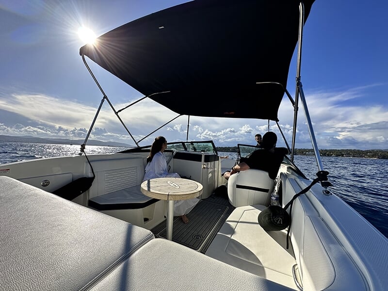 Intérieur d'un bateau avec un toit noir, une petite table et des passagers assis, profitant d'une vue sur la mer sous un ciel ensoleillé.