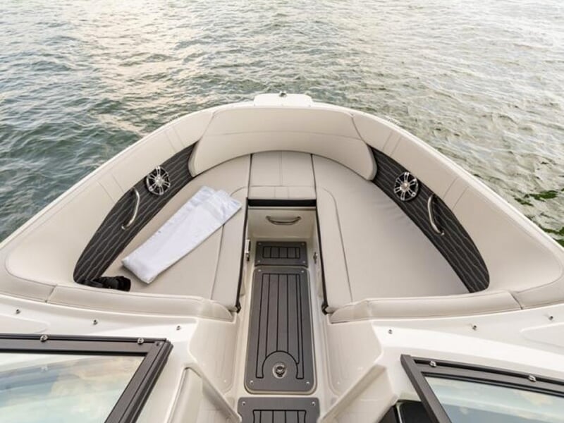 Espace avant d’un bateau de plaisance avec banquettes rembourrées beige, haut-parleurs intégrés et rangement central, entouré d’eau calme sous un ciel clair
