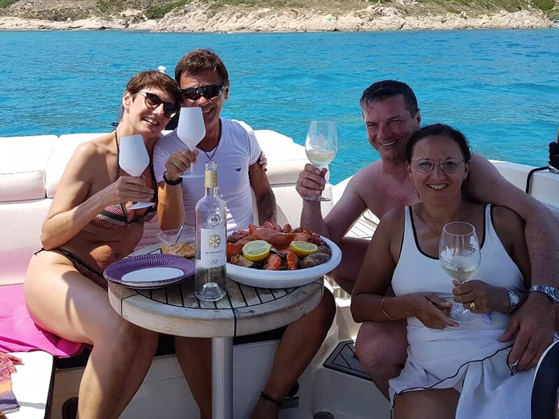 Groupe de quatre personnes souriantes partageant un repas à bord d'un bateau, avec des fruits de mer et du vin blanc sur une petite table ronde, eau turquoise et rivage en arrière-plan