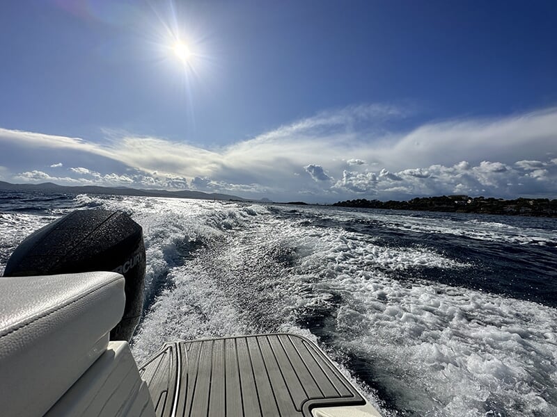 Vue arrière d'un bateau en mouvement rapide sur l'eau, montrant des vagues blanches sous un ciel ensoleillé.