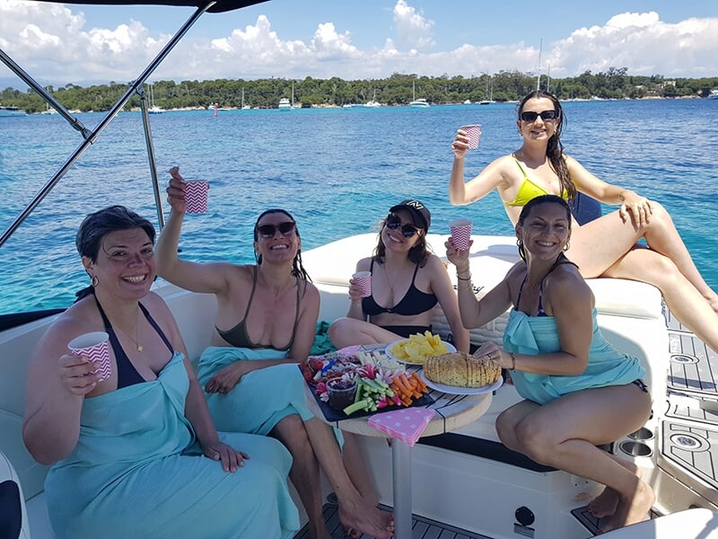 Groupe d'amies assises autour d'une table sur un bateau, partageant des snacks et levant leurs verres pour un toast.