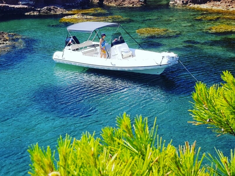 Petit bateau ancré dans une crique aux eaux transparentes, entouré de rochers et de végétation méditerranéenne.
