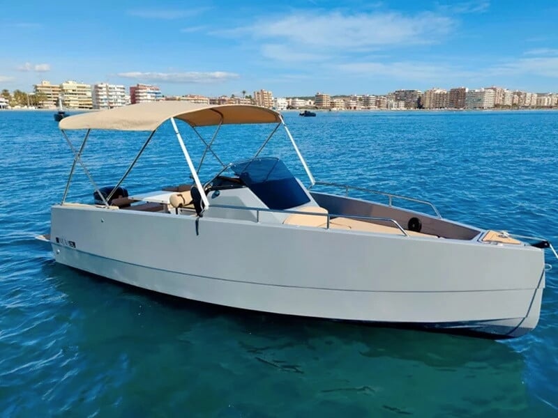 Une vedette de plaisance moderne avec une coque grise et un toit bimini beige, naviguant sur une mer calme avec des immeubles visibles en arrière-plan.