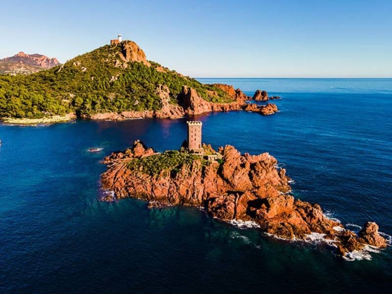 Île rocheuse avec une tour ancienne entourée d'eau bleue, végétation dense et ciel clair en arrière-plan.