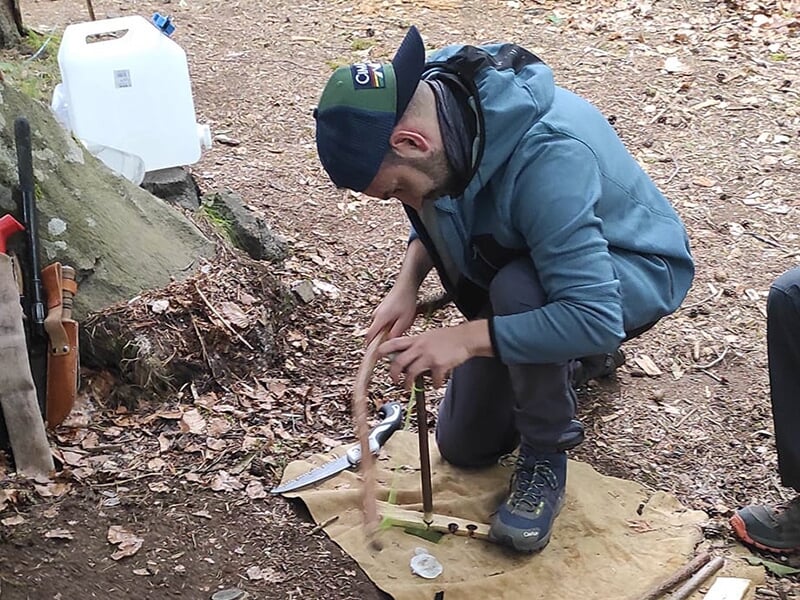 Un homme en tenue de randonnée, portant une casquette, essaye d’allumer un feu avec une technique de friction en pleine nature.