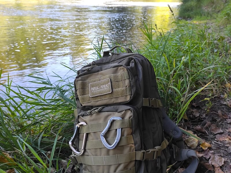 Un sac à dos militaire Mil-Tec posé sur l’herbe près d’une rivière, équipé de mousquetons et de multiples poches.