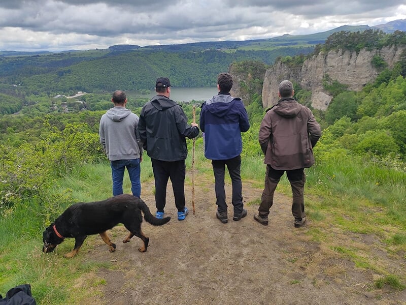 Quatre hommes debout au bord d’une falaise, accompagnés d’un chien, contemplent une vallée verdoyante avec un lac au loin.