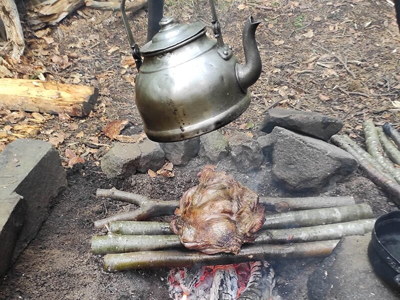 Un morceau de viande rôti sur des branches au-dessus d’un feu de camp, avec une bouilloire suspendue au-dessus.