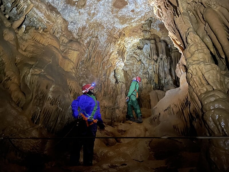 Deux spéléologues progressent à l'intérieur d'une grotte ornée de stalactites et de formations rocheuses, équipés de casques avec lampes frontales et de harnais de sécurité.