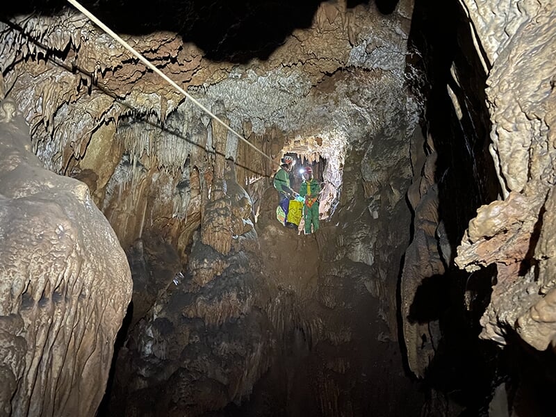 Deux explorateurs suspendus à une corde traversent un passage étroit dans une grotte, entourés de formations calcaires aux parois humides et scintillantes.