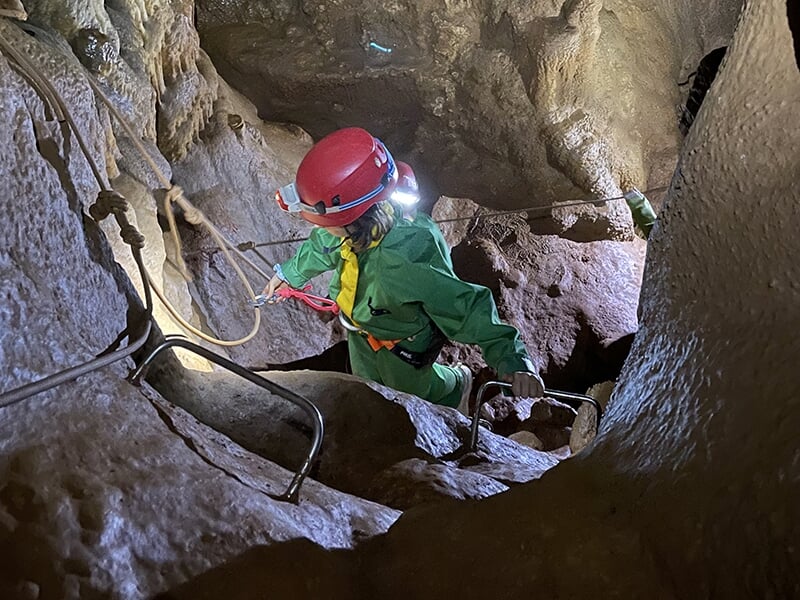 Un spéléologue en combinaison verte et casque rouge escalade une paroi rocheuse à l'aide d'étriers métalliques fixés à la grotte, s'assurant avec une longe de sécurité.