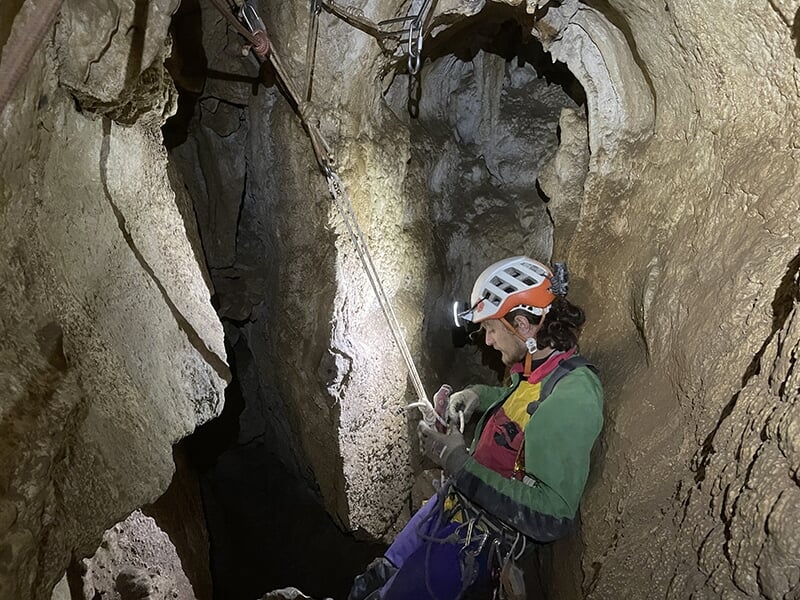 Un spéléologue équipé d’un casque et d’un harnais descend en rappel dans une grotte, éclairé par sa lampe frontale.