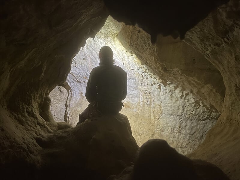 Une silhouette assise sur un rocher dans une cavité souterraine, observant l’entrée lumineuse de la grotte.