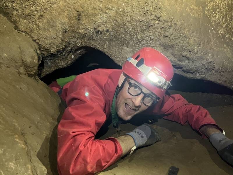 Spéléologue souriant en combinaison rouge et casque avec lampe, sortant d'un passage étroit dans la grotte du Barrage, près de Nîmes.