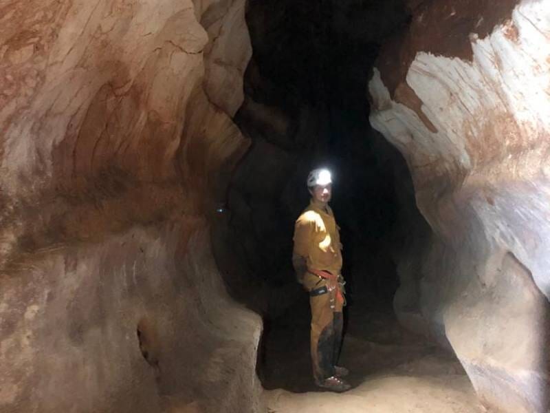 Personne en tenue de spéléologie avec casque et lampe frontale, debout dans une grande cavité rocheuse sombre.