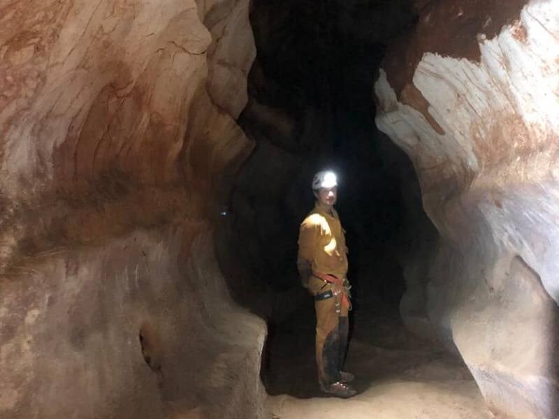 Personne en tenue de spéléologie avec casque et lampe frontale, debout dans une grande cavité rocheuse sombre.