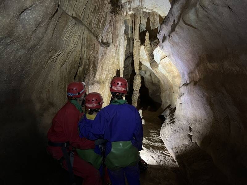 Trois spéléologues en combinaison et casque rouge explorent la grotte du Barrage, près de Nîmes, avec des stalactites éclairées par leurs lampes.