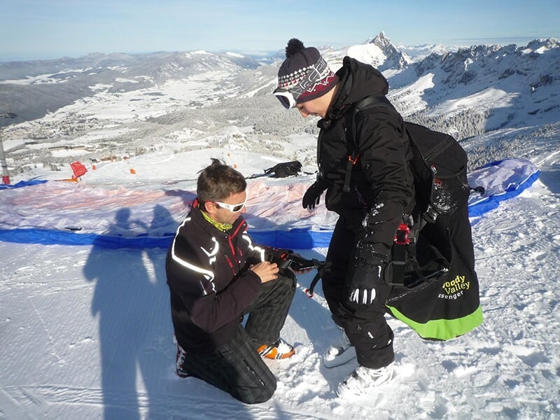 Une personne en tenue de ski est aidée par un instructeur pour se préparer au parapente sur une montagne enneigée.