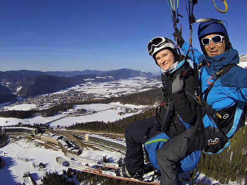 Deux personnes en tandem font du parapente au-dessus d'un paysage montagneux enneigé avec une ville en contrebas.