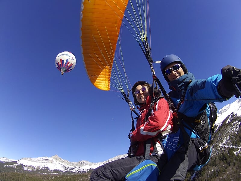 Un duo en parapente survole une vallée enneigée avec un ballon à air chaud visible en arrière-plan.