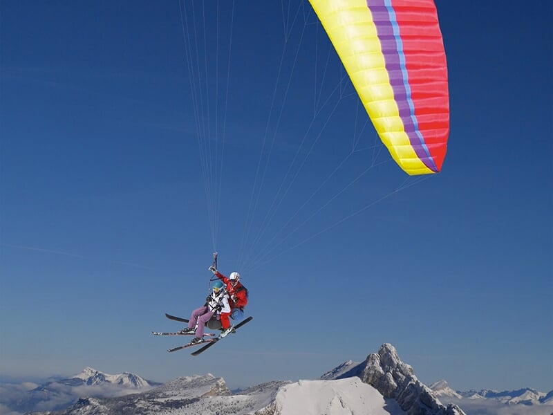 Deux personnes en parapente skis aux pieds, planent au-dessus des sommets enneigés sous une voile colorée.