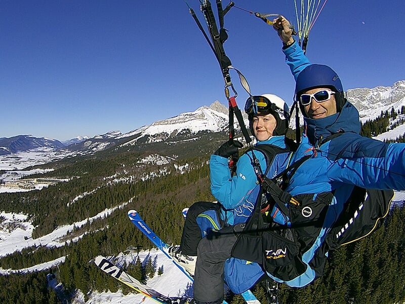 Un binôme en parapente survole une forêt enneigée, les montagnes en toile de fond.