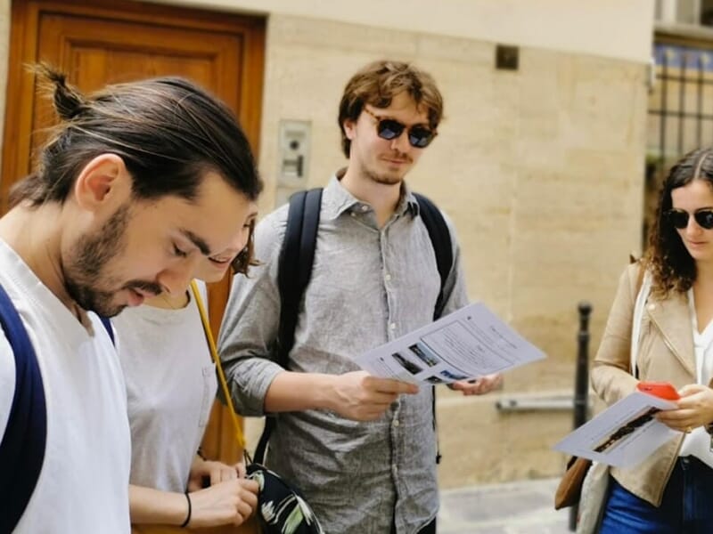 Un groupe de jeunes adultes concentrés sur une feuille de jeu de piste dans une rue de Paris.