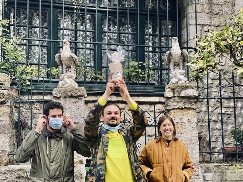Trois personnes devant une façade en pierre, une d'elles tenant un trophée emballé dans du plastique.
