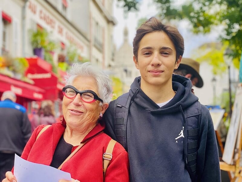 Une femme âgée souriante avec des lunettes et un jeune garçon en sweat noir dans une rue animée.