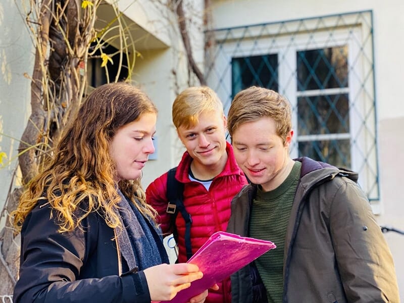 Trois jeunes adultes regardent un document rose à l’extérieur, l’une d’elles semble lire à haute voix.
