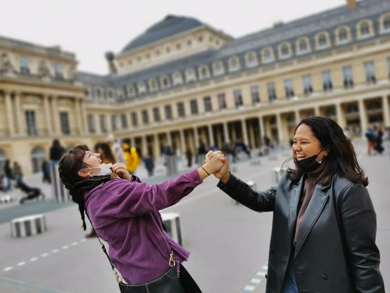 Deux amies rient et se tiennent les mains devant les colonnes de Buren au Palais Royal à Paris.