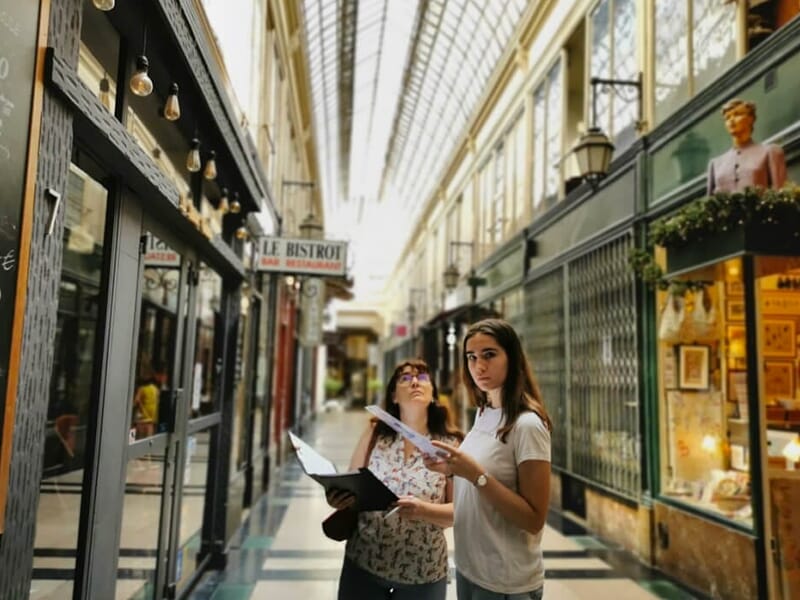 Deux femmes observent un plan dans une galerie marchande parisienne couverte, entourées de boutiques anciennes.